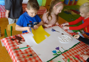 Piątka dzieci przygotowuje plakat, wycinają papierowe elementy z wycinanek, naklejają kwiaty.
